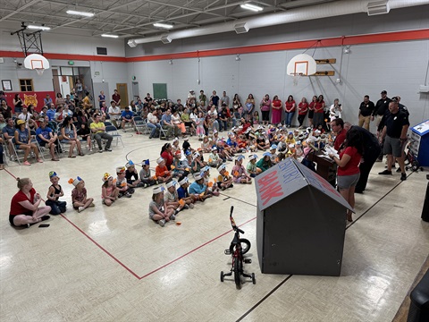 Safety Town Graduation with Kindergartners sitting in front of LAPD presenters and Safety Town supplies, with parents in the background in seats watching the celebration
