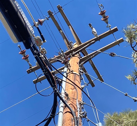 A view of a utility pole from the ground looking toward the blue sky. Several utility lines intersect at the top of the pole.
